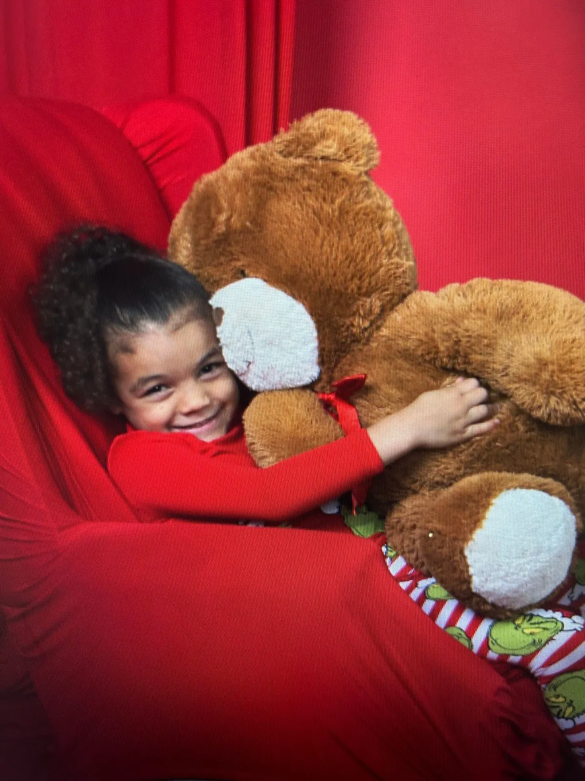 A little girl in red pajamas hugging a giant teddy bear on a red Christmas set