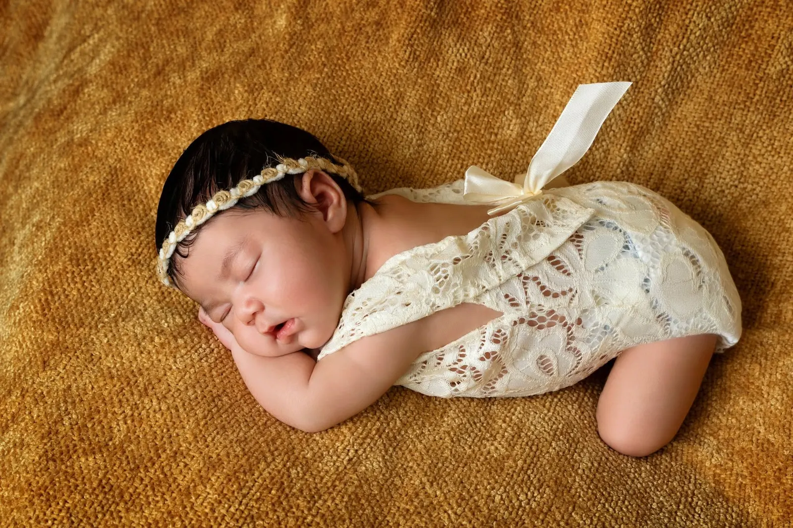 A newborn in a lace romper and headband asleep on a golden blanket