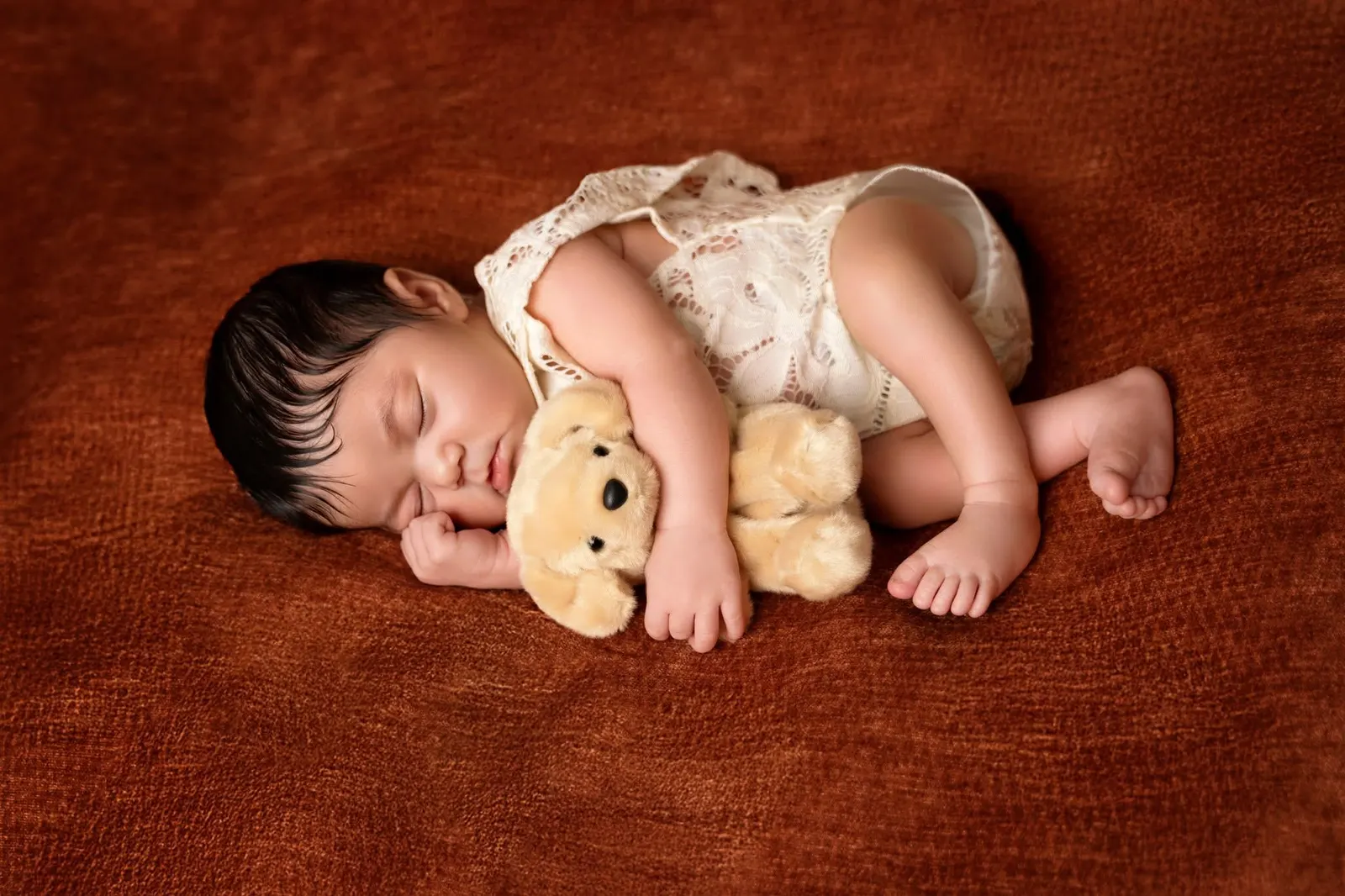 A sleeping newborn curled up with a small teddy bear on a rust-colored blanket