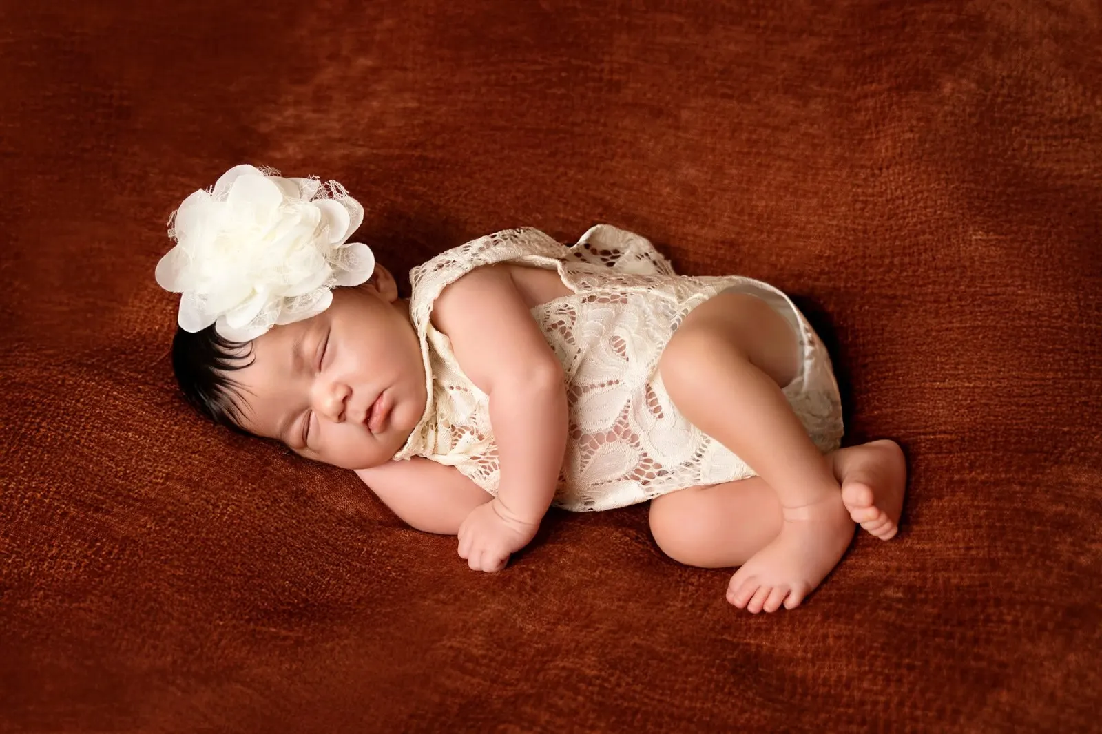 A newborn with a cream flower headband sleeping on a rust blanket