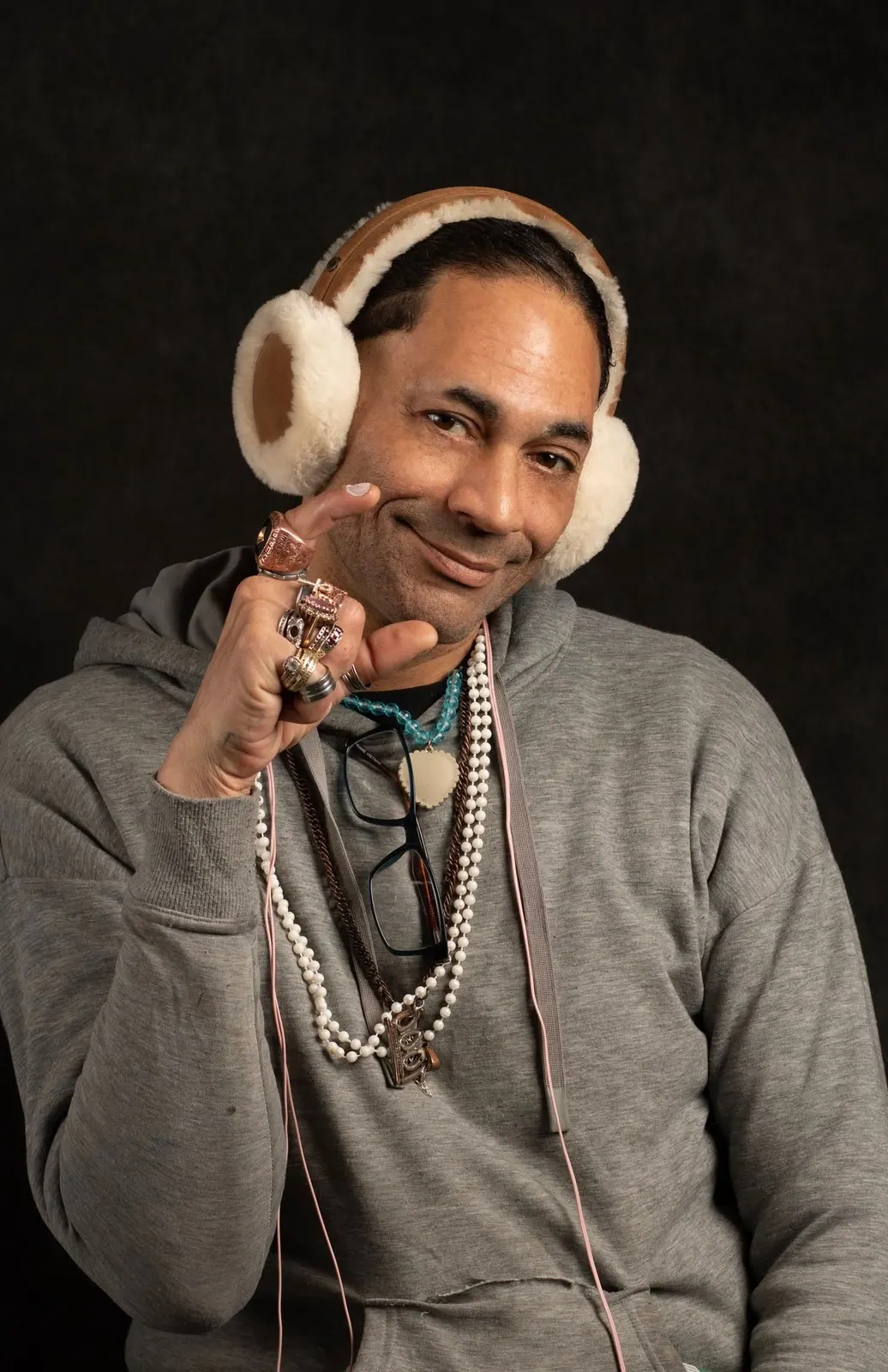 A man in cream earmuffs and pearls smiling on a dark backdrop