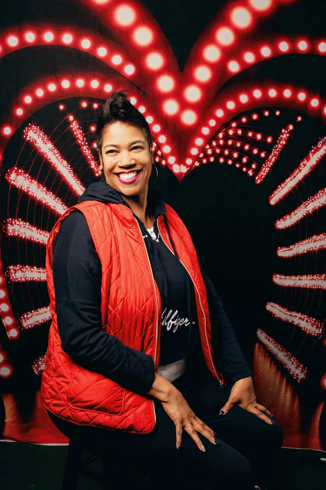 A smiling woman in a red puffer vest posed in front of a heart of marquee lights