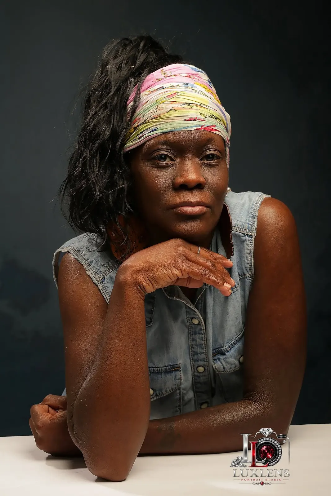 A woman in a denim vest and headwrap resting her chin on her hand in a studio portrait