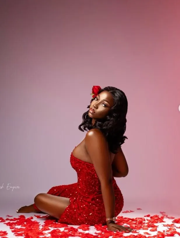 A woman in a sparkling red dress seated among rose petals on a blush backdrop